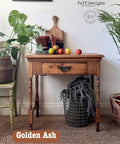 A rustic wooden table holds fruits, cutting boards, and a candle. It's flanked by green plants in woven baskets. A vintage chair and textured rug complete the cozy setting. Text reads "Golden Ash" and "Faff designs.