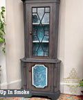A vintage corner cabinet with a gray finish and blue interior, featuring geometric glass panels. It's decorated with potted plants on either side. The text "Up In Smoke" is visible at the bottom left.