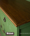 Close-up of a wooden cabinet with a green painted base and a dark walnut-stained top. The surface is glossy, showing a smooth texture. Three decorative metal handles are visible. The words "Walnut" and "Gracie's House Painted" appear on the image.