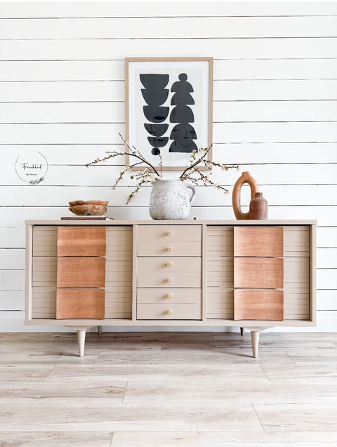 A mid-century modern sideboard cabinet with a light beige wood finish and contrasting warm brown drawer fronts, styled with home decor against a white shiplap wall.