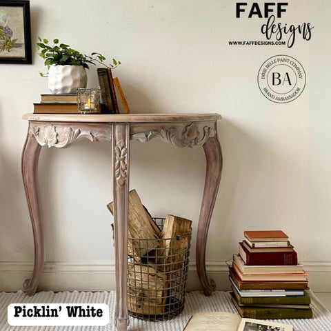 A vintage-style curved table with ornate legs is against a light wall. It holds a plant, books, and a lit candle. A wire basket with wood logs and scattered books surround the table. Text reads "Picklin' White" and "FAFF designs.