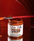 A jar of Dixie Belle Prime Start paint with an orange paint-dipped brush resting on top. Some paint drips down the jar, and the background features layered, curved, reddish-brown panels.