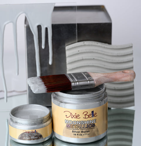 Jars of 'Dixie Belle Moonshine Metallics Silver Bullet' paint with a brush and decorative metal wall art in the background.