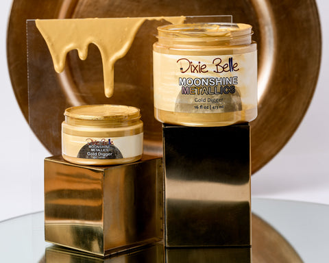 Two jars of 'Moonshine Metallic Gold Digger 'by Dixie Belle on a reflective surface with a metallic background.