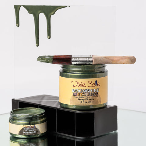 Jars of 'Dixie Belle Moonshine Metallics' with a paintbrush on a glass surface.