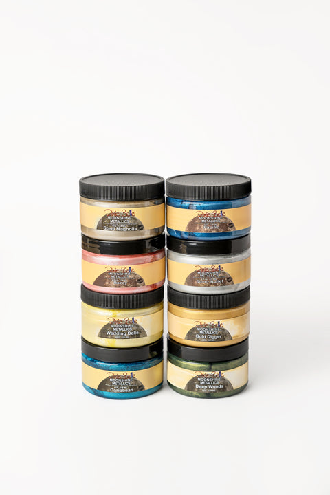 Eight jars of Dixie Belle Moonshine Metallics paint are stacked in two columns. Each jar has a black lid and a yellow label, with different colors visible through the containers.