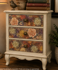 A vintage-style three-drawer nightstand with a distressed cream finish and floral drawer fronts, shown in a styled room setting next to a lamp and potted plant.