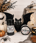 Two product jars displayed on a white wood surface with seasonal props including a black bat cutout, spider decorations, a small pumpkin, lace, and a candle. The jar on the left reads 'Velvet Touch Black' and the jar on the right reads 'Stick With Me'.