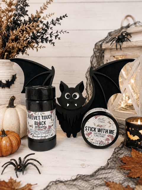 Two product jars displayed on a white wood surface with seasonal props including a black bat cutout, spider decorations, a small pumpkin, lace, and a candle. The jar on the left reads 'Velvet Touch Black' and the jar on the right reads 'Stick With Me'.