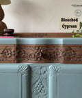 A vintage sideboard with ornate wood carvings is painted in light blue. It features two large cabinet doors with wooden handles. Potted plants and a table lamp with a woven base are placed on and around it. The setting includes a woven rug.