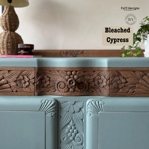 A vintage sideboard with ornate wood carvings is painted in light blue. It features two large cabinet doors with wooden handles. Potted plants and a table lamp with a woven base are placed on and around it. The setting includes a woven rug.