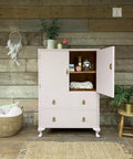 a pink cabinet painted in all in one paint with top right door open revealing a blanket and some books and vases with a pink flower.  On top is a pitcher with greenery and two candles sit next to that.  on wall is a dream catcher, to the left of cabinet is a wicker basket with pillows and to the right is a chair with a plant