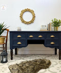 A side table painted with all in one paint in a dark blue with gold pulls for hardware, staged with a plant in a wicker basket to the left, gold mirror on wall and plant and books with a coffee mug on top