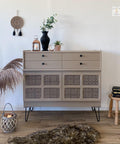 side table painted in a tan all in one paint, with four drawers on top and bottom has 8 squares that are inlays with decoupage. to the left is a container with pampas grass, on top is a candle and vases with plants, to the right is a stool with books and in front is a furry rug and a basket with a candle