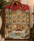 A decorative cutting board with a vintage storefront building decoupage paper, shown in a styled holiday scene with a candle, vase, evergreen branches, and small winter figures in front of the shop. The ornament has a red top section, tan and gray building facade, warm yellow window lights, and a snowy ground scene.