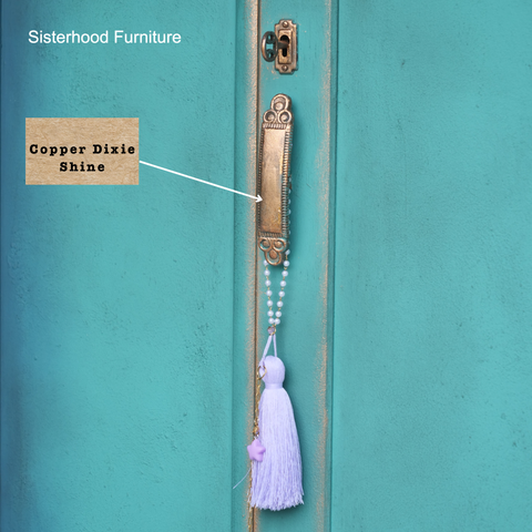 Decorative door handle with tassel on a teal door, featuring 'Sisterhood Furniture' and 'Copper Dixie Shine' text.
