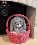 A gray plush bunny with a plaid bow tie sits inside a bright pink woven basket placed in front of a stone fireplace. The basket has a visible 'Born Again Refinishing Studio' logo on the left side.