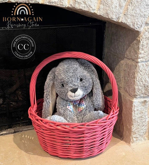 A gray plush bunny with a plaid bow tie sits inside a bright pink woven basket placed in front of a stone fireplace. The basket has a visible 'Born Again Refinishing Studio' logo on the left side.