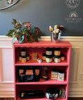 A pink wooden three-shelf storage cabinet in a home setting, styled with various craft supplies including jars, brushes, a potted plant, and a tool bag.