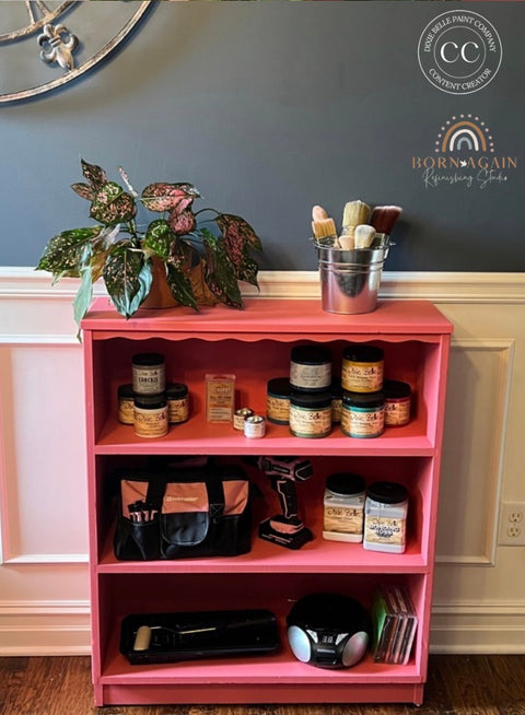 A pink wooden three-shelf storage cabinet in a home setting, styled with various craft supplies including jars, brushes, a potted plant, and a tool bag.