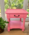 A bright pink wooden end table with a single drawer and lower shelf, styled in a sunroom next to a potted plant and a wicker chair.