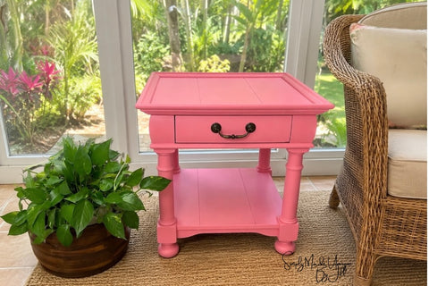 A bright pink wooden end table with a single drawer and lower shelf, styled in a sunroom next to a potted plant and a wicker chair.