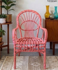 A coral pink rattan armchair with a high curved backrest, shown in a styled room setting on a patterned rug with wooden furniture and plants in the background.