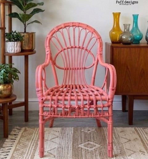 A coral pink rattan armchair with a high curved backrest, shown in a styled room setting on a patterned rug with wooden furniture and plants in the background.