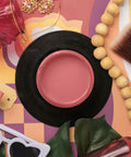 A styled flat-lay featuring a round pink paint jar on a black circular base, surrounded by colorful props including a paintbrush, wooden beads, a red glass bottle, sunglasses, and a paint palette on a patterned background.