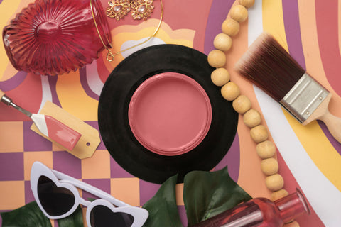 A styled flat-lay featuring a round pink paint jar on a black circular base, surrounded by colorful props including a paintbrush, wooden beads, a red glass bottle, sunglasses, and a paint palette on a patterned background.