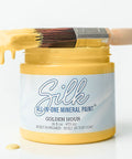 A jar of Silk Golden Hour mineral paint with yellow paint dripping from a paintbrush resting across the open jar, set against a clean white background.