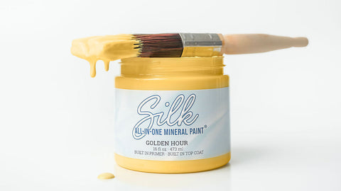 A jar of Silk Golden Hour mineral paint with yellow paint dripping from a paintbrush resting across the open jar, set against a clean white background.
