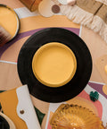 A styled flat-lay featuring a round yellow paint jar with a paintbrush, a black-rimmed yellow paint jar lid, a palette knife, a yellow wallet, sunglasses, a goldish yellow glass jar, and tropical leaves on a colorful abstract background.