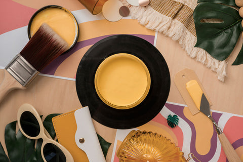 A styled flat-lay featuring a round yellow paint jar with a paintbrush, a black-rimmed yellow paint jar lid, a palette knife, a yellow wallet, sunglasses, a goldish yellow glass jar, and tropical leaves on a colorful abstract background.