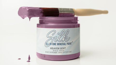 A jar of Silk All-In-One Mineral Paint in the shade Heaven Sent, featuring a lavender-purple paintbrush resting across the open jar on a clean white background.