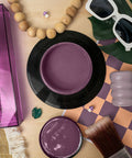 A styled flat-lay featuring a purple paint jar with a metal palette tool on a purple box, a round jar lid with purple paint inside, an open jar, a brush, sunglasses, wooden beads, green leaves, and a checkered mat on a light wood surface.