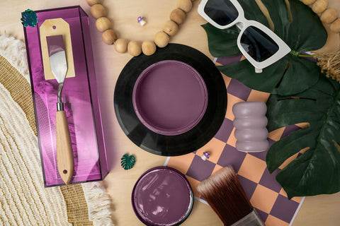 A styled flat-lay featuring a purple paint jar with a metal palette tool on a purple box, a round jar lid with purple paint inside, an open jar, a brush, sunglasses, wooden beads, green leaves, and a checkered mat on a light wood surface.
