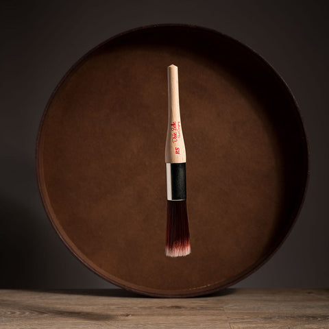 Paint brush with wooden handle and red bristle head floating in front of a dark brown circular platform.