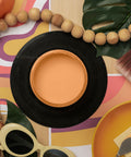 A styled flat-lay featuring a round orange paint jar, surrounded by tropical leaves, wooden beads, a paintbrush, and colorful patterned paper.