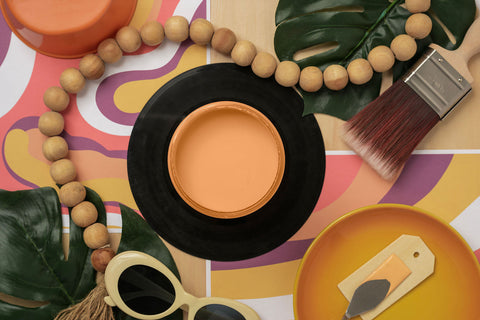 A styled flat-lay featuring a round orange paint jar, surrounded by tropical leaves, wooden beads, a paintbrush, and colorful patterned paper.
