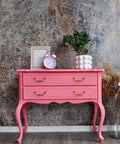 A bright pink vintage-style console table with two drawers and curved legs, styled with a pink alarm clock, a potted plant, and a picture frame against a textured gray wall.