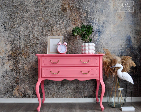 A bright pink vintage-style console table with two drawers and curved legs, styled with a pink alarm clock, a potted plant, and a picture frame against a textured gray wall.