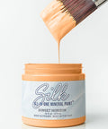 A jar of Silk All-In-One Mineral Paint in Sunset Horizon, with a paintbrush dipped in the orange paint dripping into the jar on a white background.