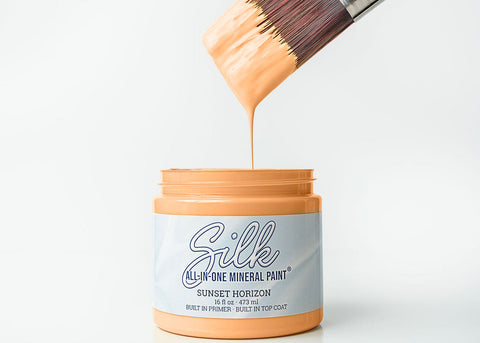A jar of Silk All-In-One Mineral Paint in Sunset Horizon, with a paintbrush dipped in the orange paint dripping into the jar on a white background.
