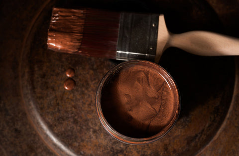 Close-up of a can of copper metallic paint with a brush on a rustic metal surface.
