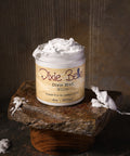 A jar of Dixie Belle Dixie Mud White, 8 oz (237 mL), sits on a stone slab with a wood base. The label reads "Straight from the swamps of Dixie." The background is brown and textured.