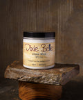 A jar of Dixie Belle Dixie Mud White sits on a stone slab. The jar is open, showing white mud paste with some spread on the stone and a tool. The label reads “Straight from the swamps of Dixie.”.