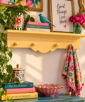 A styled interior scene with a yellow floating shelf holding framed art, books, a blue bird figurine, flowers, and a patterned towel. A turquoise console table below holds a pink scalloped bowl and stacked books, with a green plant in the foreground against a white wall.