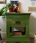 Green wooden side table with a drawer, books, and potted plants in a room setting. Labeled "English Ivy blended with Florida Orange."
