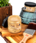 Dixie Belle Gold Shimmer Glaze jar with a brush on a wooden surface, surrounded by decorative items.